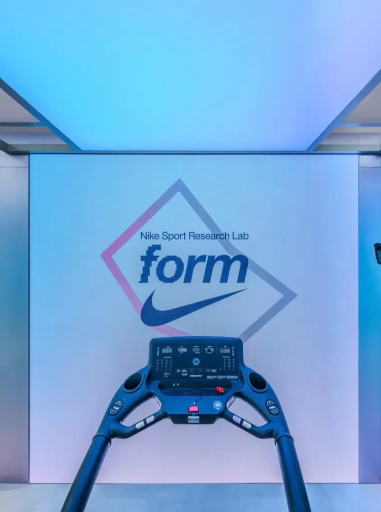 Front-facing view of a treadmill console inside Nike Sports Research Lab pop-up at Oxford Circus, set against a softly lit gradient wall displaying the NSRL “form” graphic and Swoosh branding in a minimal, clinical environment