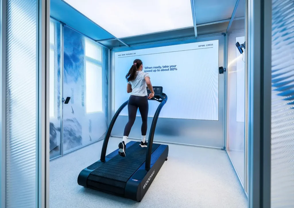 Visitor running on a curved treadmill inside Nike Sports Research Lab pop-up at Oxford Circus, surrounded by minimalist metallic interiors and a digital instruction screen guiding performance testing speed adjustments
