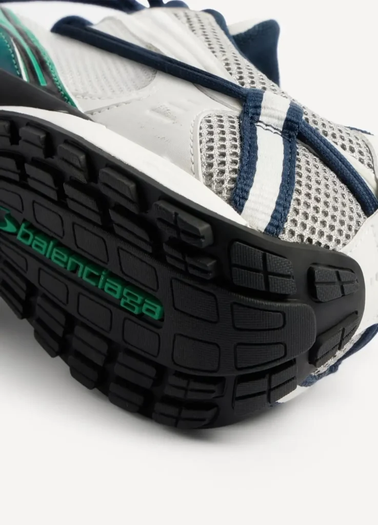 Close-up of the Balenciaga Radar Shoe outsole featuring a segmented black rubber tread with grid-like traction pattern and embedded green Balenciaga branding; upper shows breathable white mesh with perforated texture, navy lace system wrapping across the foot, and subtle layered paneling that highlights the shoe’s lightweight, flexible construction