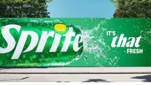 Wide outdoor billboard featuring a vibrant green Sprite campaign visual, showing a close-up of a Sprite can bursting with water splash and condensation, alongside bold white text reading “It’s that fresh,” set against a clean urban streetscape with trees and blue sky, emphasizing refreshment, scale, and high-impact branding