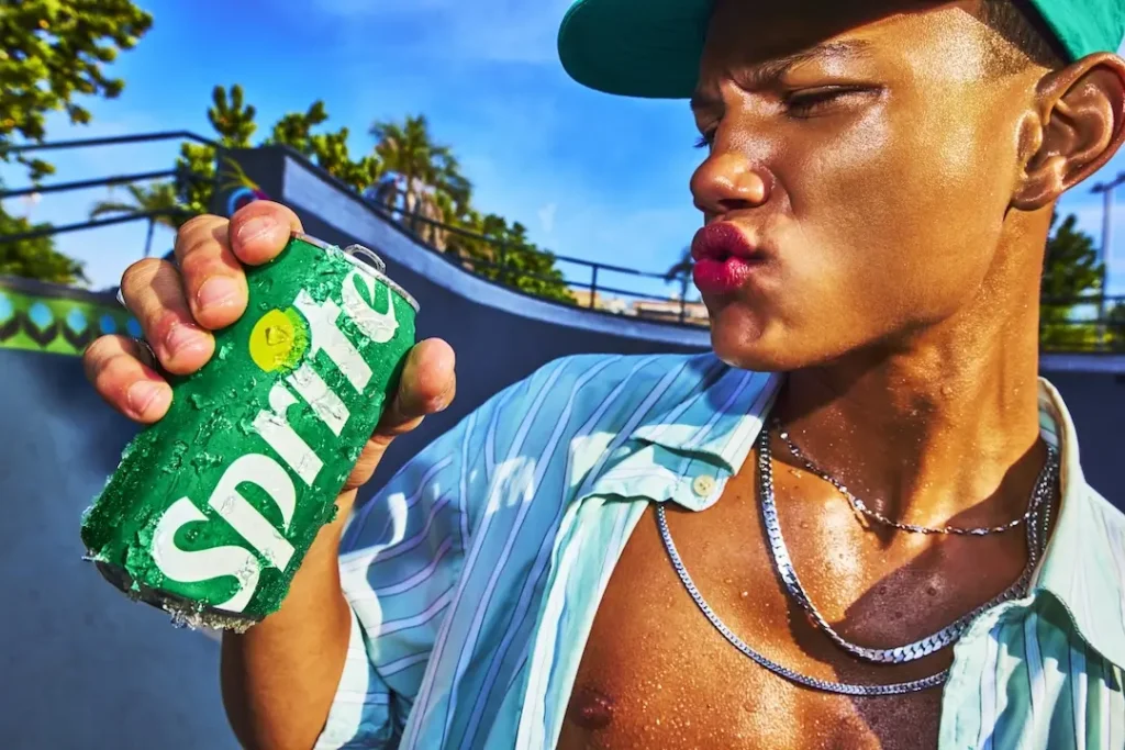Close-up of a person in sunlit outdoor setting holding an ice-cold Sprite can covered in condensation, wearing a green cap, open striped shirt, and silver chain, puckering their lips as if reacting to the drink’s sharp citrus taste, with a skatepark and palm trees blurred in the background, emphasizing freshness, heat, and urban lifestyle energy