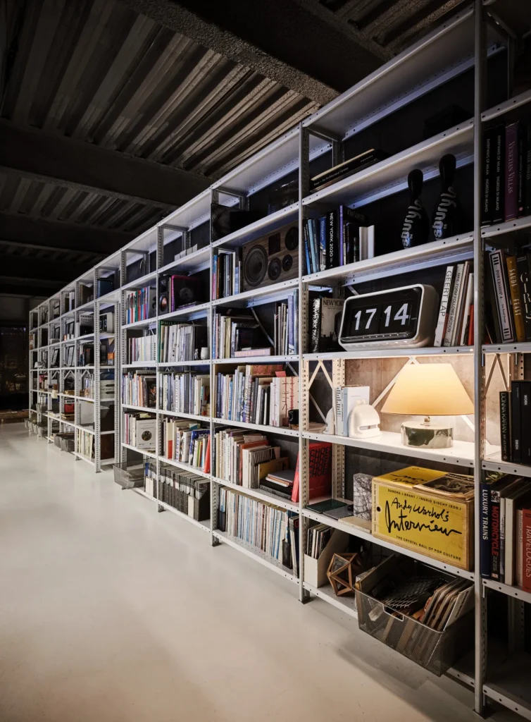 A long wall of industrial metal shelving filled with books, design objects, and speakers, softly lit by integrated lighting, forming a curated library within a contemporary studio space