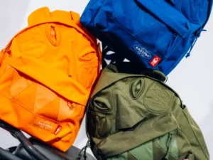 Three colorful Eastpak backpacks from the Walter Van Beirendonck collaboration featuring sculpted monster-like faces in orange, blue, and olive green with stitched eye and mouth details
