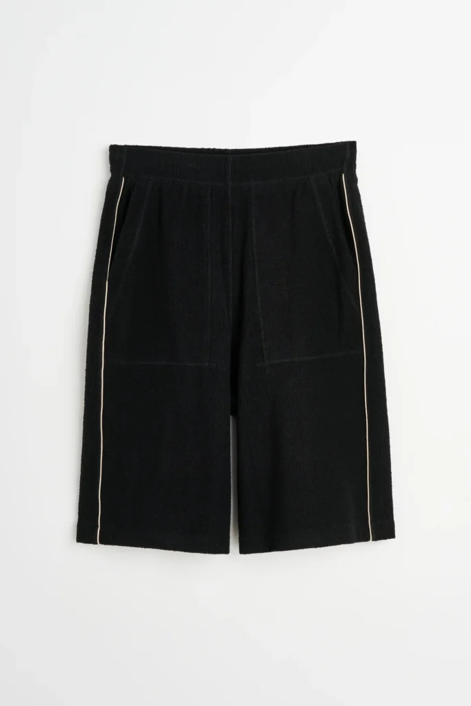 Black textured shorts from the Wales Bonner Spring Summer 2026 collection featuring a relaxed fit, elastic waistband, front seam paneling, and thin contrast piping along the side seams against a neutral background