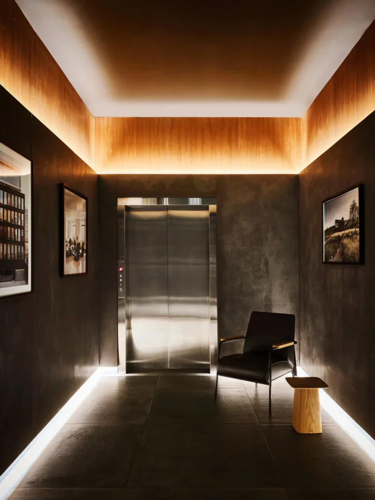 Alt Text (Mid-Length, Editorial-Ready) A warm, minimalist elevator lobby with dark textured walls, a softly backlit wood ceiling, stainless steel elevator doors, and a single lounge chair with a small wooden side table creating a quiet, refined atmosphere