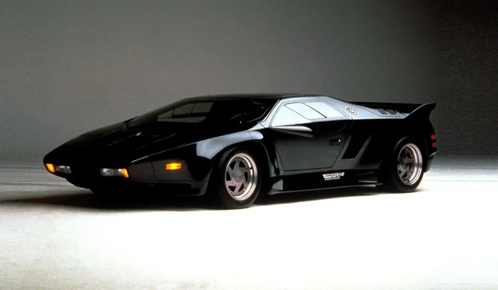 Black Vector W8 Twin Turbo American supercar in side profile, highlighting its sharp wedge body, wide rear wing, and futuristic aerospace-inspired design. Alt text: Top-down view of a black Vector W8 Twin Turbo supercar driving on a road, highlighting its angular wedge shape, central hood vents, and aerodynamic fighter-jet inspired design. Alt text: Red Vector W8 Twin Turbo supercar with scissor doors and rear engine cover raised, photographed in a mountainous desert landscape at dusk, showcasing the dramatic wedge-shaped American supercar design. Alt text: Vector W8 interior cockpit featuring digital dashboard display, aircraft-style toggle switches, and a futuristic steering wheel with teal accents, emphasizing the supercar’s aerospace-inspired design