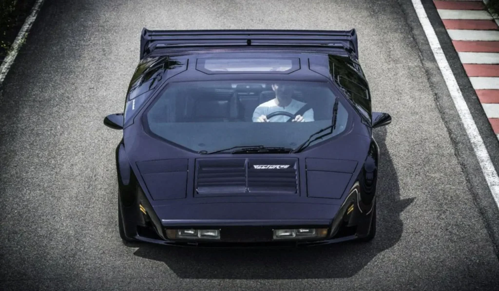 Top-down view of a black Vector W8 Twin Turbo supercar driving on a road, highlighting its angular wedge shape, central hood vents, and aerodynamic fighter-jet inspired design