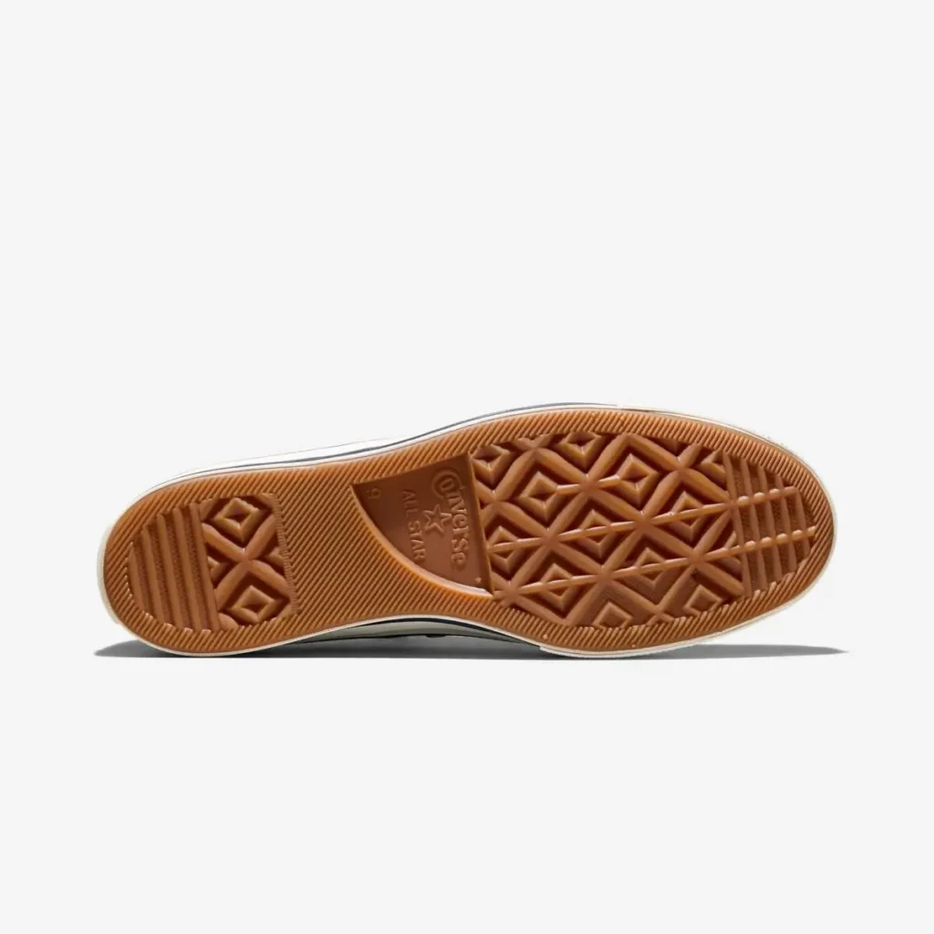 Outsole view of Converse Chuck 70 shoe featuring a gum rubber sole with classic diamond tread pattern, textured grip zones, and central Converse All Star branding; the warm brown outsole contrasts with the upper, consistent with a Noah collaboration’s vintage-inspired finish