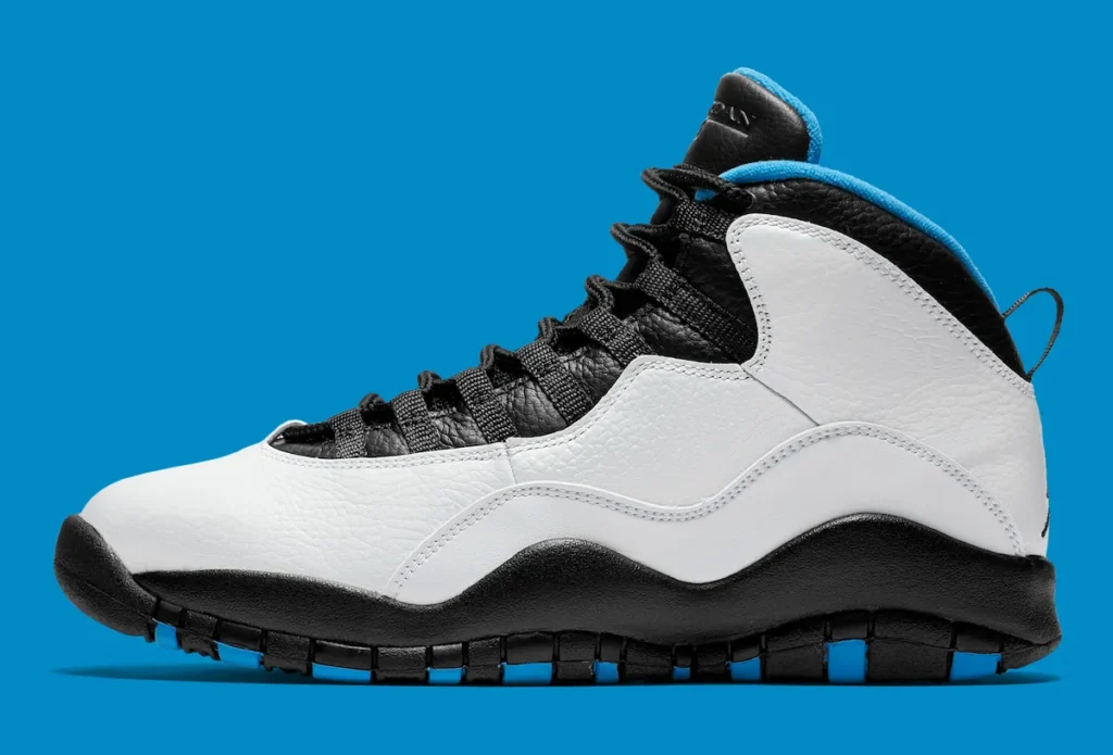 Side profile of the Air Jordan 10 “Powder Blue” shoe against a bright blue background, featuring a white tumbled leather upper, black textured leather eyestay and tongue, black rope laces, a black midsole, and subtle powder blue accents along the collar and outsole