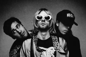 Kurt Cobain wearing white oval sunglasses standing between bandmates Dave Grohl and Krist Novoselic in a black-and-white Nirvana portrait