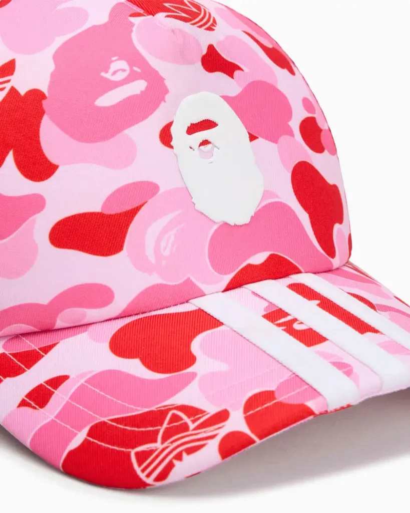 Close-up of adidas Originals x BAPE trucker cap featuring pink ABC camouflage fabric, white BAPE ape head logo patch, and adidas three stripes across the curved visor