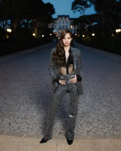 Lisa stands centered on a gravel pathway at dusk, wearing a sharply tailored pinstripe suit layered over a sheer black top, with a softly lit villa and trees framing the background in a cinematic evening setting