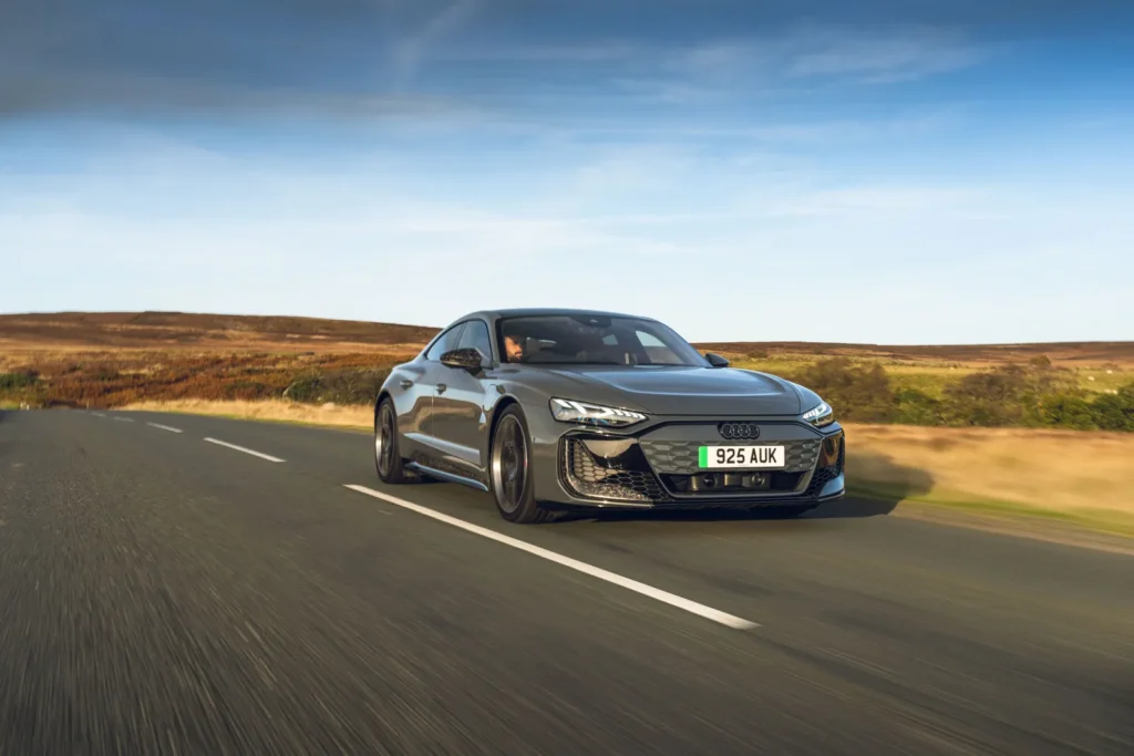 Audi e-tron GT RS Performance driving at speed on an open road, highlighting its aggressive front fascia, low-slung stance, aerodynamic lines, and dynamic motion through a sweeping countryside landscape