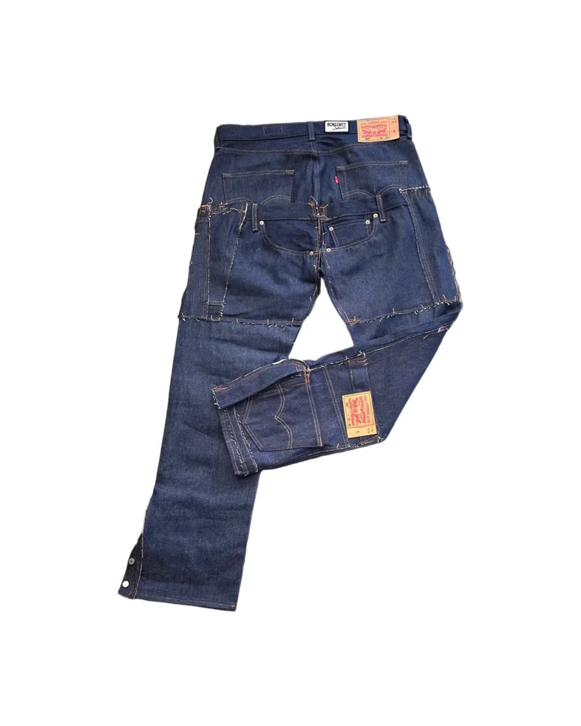 RCNSTRCT Studio Raw Denim Multi Pocket Pant in dark indigo raw denim shown in angled back view, featuring layered patchwork panels, reinforced utility pockets, contrast stitching, and structured streetwear silhouette