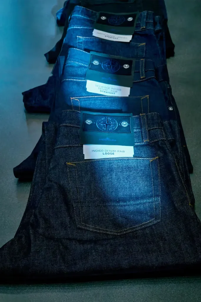 Folded stacks of raw indigo denim jeans from Stone Island Denim Research, displaying back pockets, contrast stitching, and branded compass patch labels indicating different selvedge indigo fits and washes