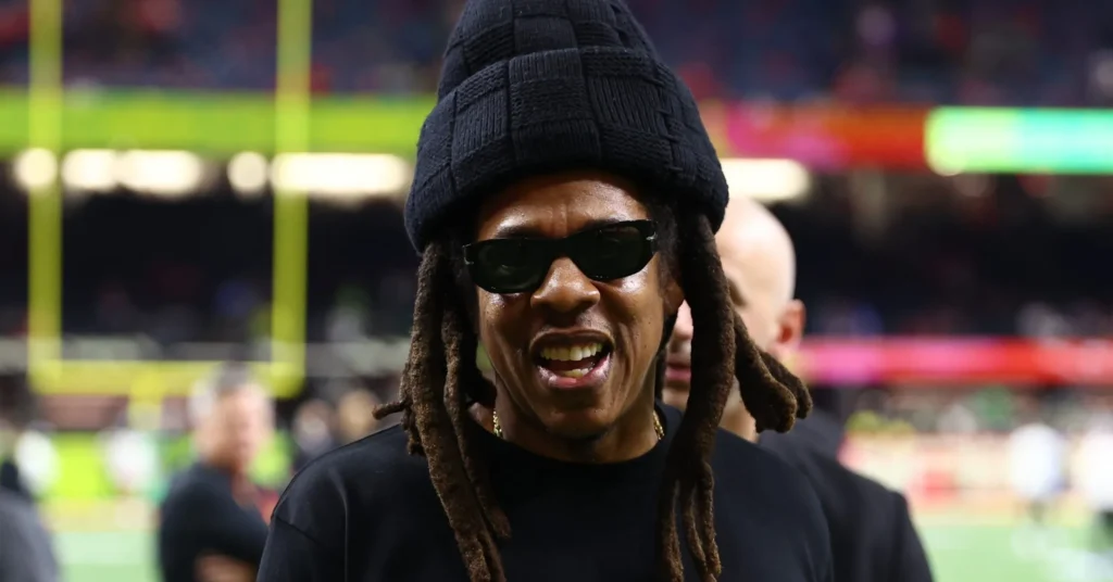 Jay-Z wearing a black beanie and sunglasses, smiling on the field inside a stadium with blurred crowd and goalpost in the background