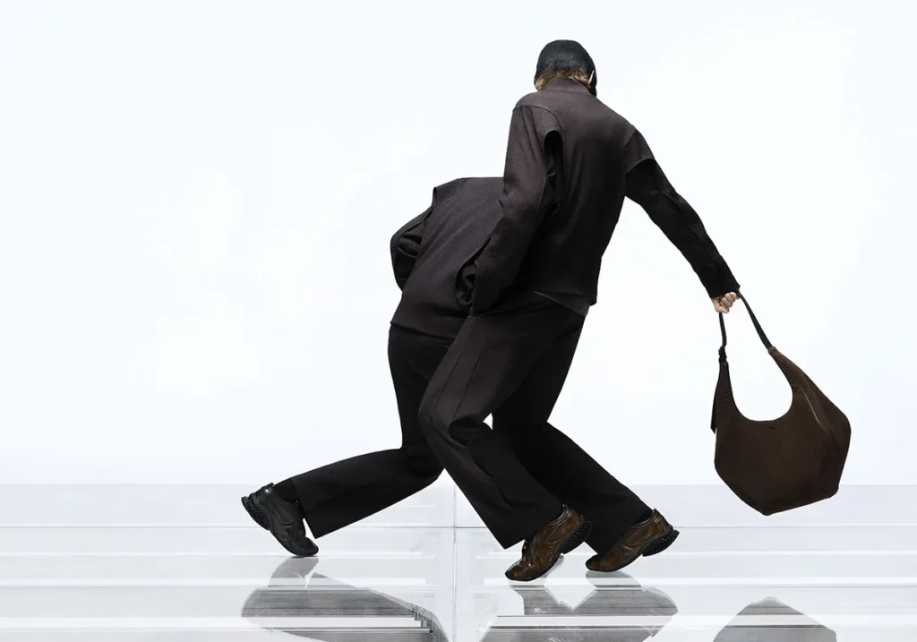 Two models in motion wearing dark, tailored technical outfits, captured mid-stride on a reflective platform, with one carrying a large brown hobo-style bag and both styled in PUMA x J.L-A.L footwear against a minimal studio backdrop