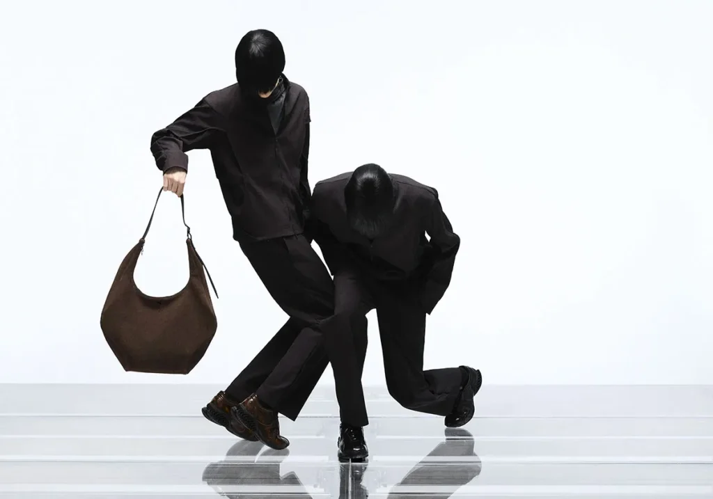 Two models in coordinated dark technical outfits captured in a dynamic, sculptural pose, with one holding a large brown hobo-style bag, both wearing PUMA x J.L-A.L footwear on a reflective platform against a minimal studio background