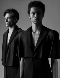 Two male fashion models posed in a minimalist studio portrait wearing sculptural black artisanal menswear garments with draped sleeves, showcasing contemporary designer tailoring