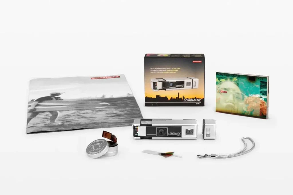 Complete Lomomatic 110 Camera & Flash Metal silver kit displayed on a neutral background, including the compact camera, detachable flash, film cartridge, carrying strap, packaging box, printed materials, and branded envelope with monochrome surf imagery