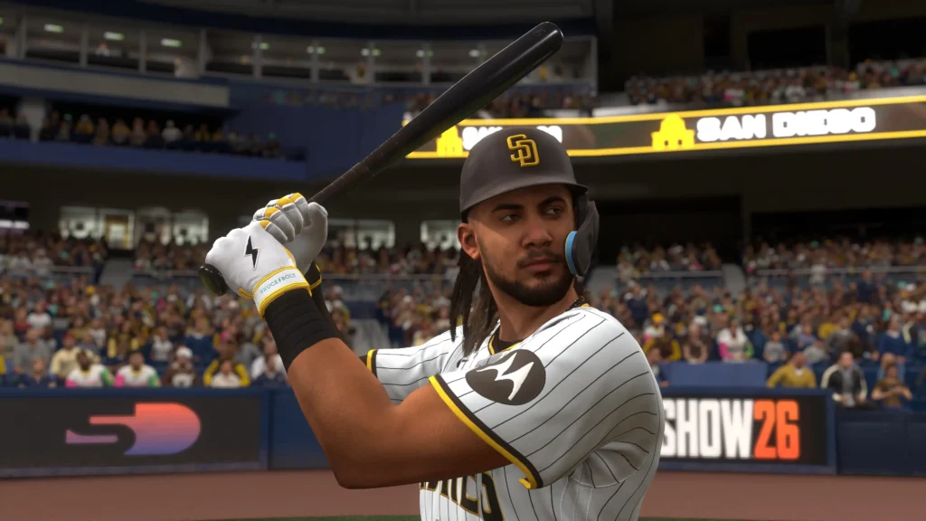 San Diego Padres batter at the plate holding a bat in a ready stance, wearing a helmet and batting gloves, with a packed stadium and MLB The Show 26 signage visible in the background
