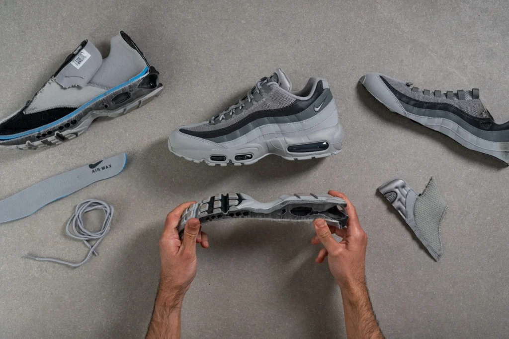 Top-down view of a deconstructed Nike Air Max 95, with multiple components—upper panels, sole unit, laces, and insole—laid out across a neutral surface, while hands hold and flex the segmented midsole to reveal its cushioning structure
