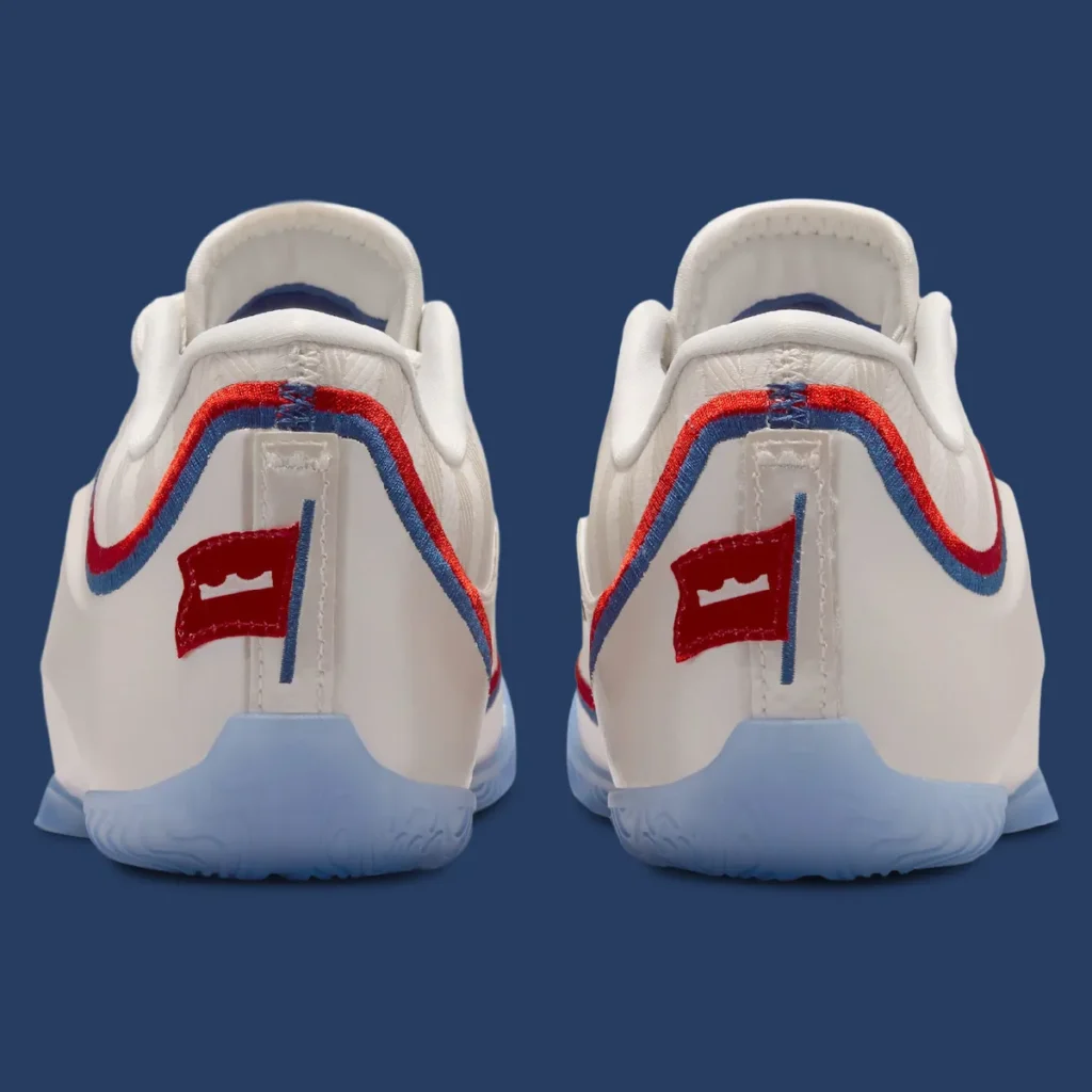 Rear view of the Nike LeBron 23 “Old Glory” showing dual heels in Pale Ivory with red and navy trim, featuring a stitched crown-and-flag emblem at the back tab, textured white upper, and translucent icy blue outsole set against a solid blue background
