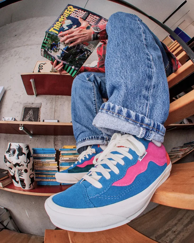 Lifestyle shot of Parra x Vans OTW Old Skool 36 sneakers worn with cuffed blue jeans, featuring blue suede panels with pink wavy overlays and white laces, captured from a low angle as the wearer sits on wooden stairs reading a Thrasher magazine in a cozy, bookshelf-lined interior
