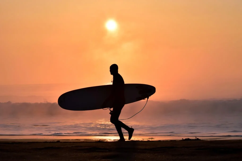 Silhouette of a surfer carrying a surfboard along the shoreline at sunset, with the sun glowing above the horizon and waves rolling in the background