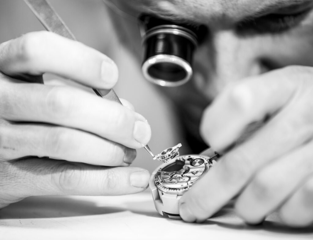 Black-and-white close-up of a watchmaker using precision tools under a loupe to assemble a mechanical watch movement by hand