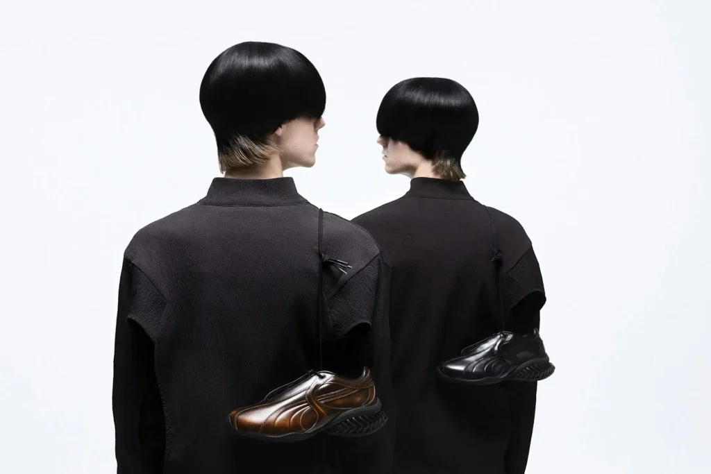 Two models seen from behind in minimalist black technical garments, each carrying a single PUMA x J.L-A.L sneaker slung over the shoulder—one in ombré brown and one in glossy black—set against a clean studio background