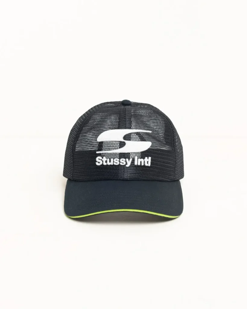 Black mesh trucker cap with curved brim, neon green edge detail, and white “Stüssy Intl” logo embroidered on the front panel