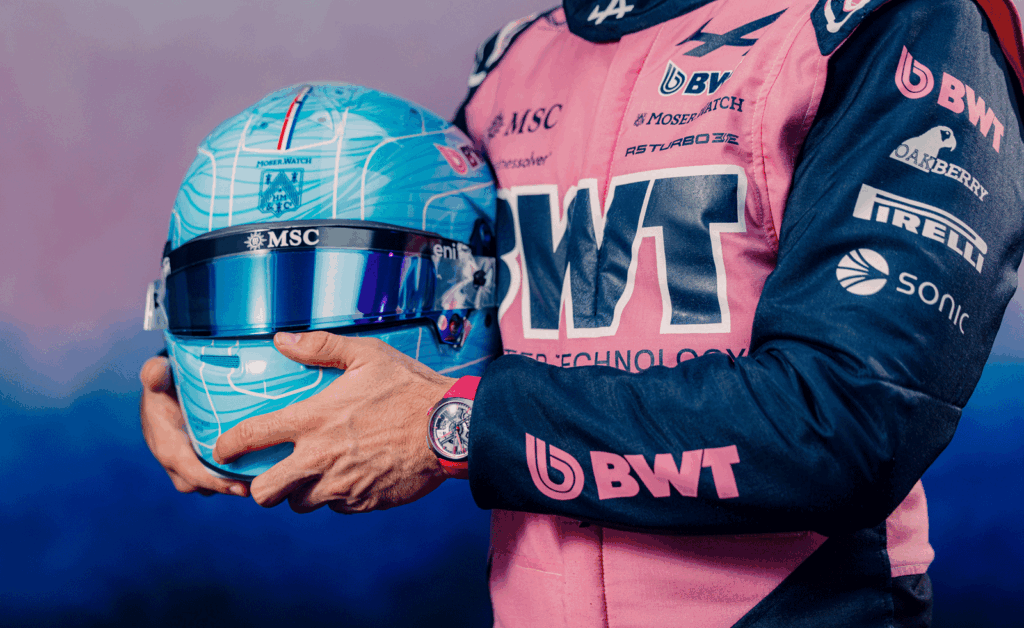 Close-up of a Formula 1 driver in a pink and black BWT Alpine racing suit holding a blue helmet with reflective visor, while wearing a H. Moser Streamliner watch on the wrist