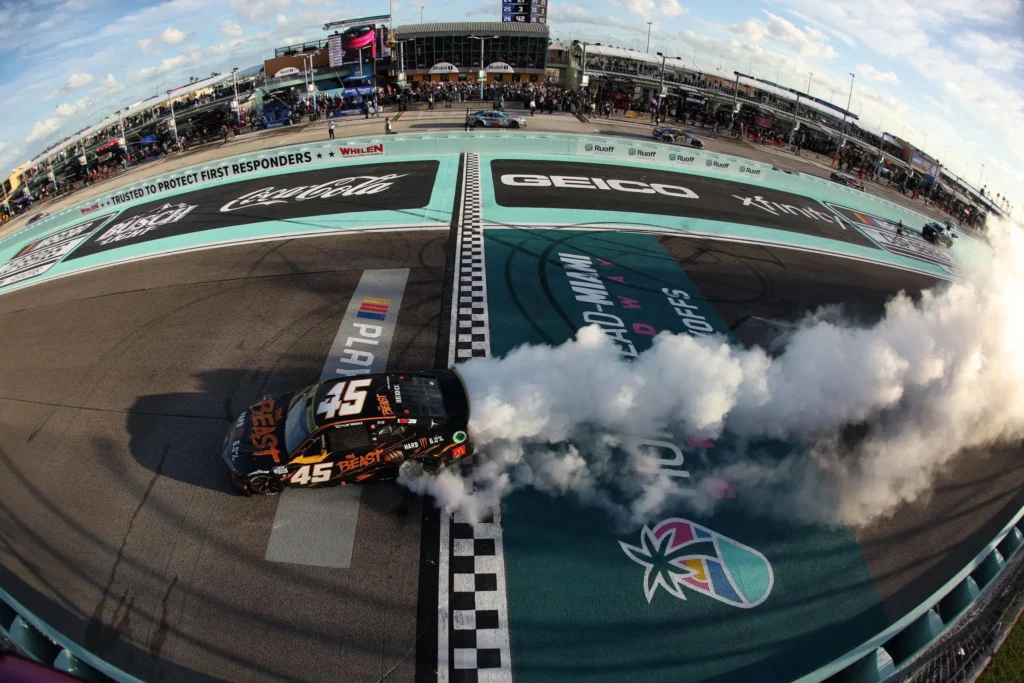 Tyler Reddick’s No. 45 23XI Racing Toyota performs a smoky burnout across the finish line after a NASCAR victory, with tire smoke billowing over the track and grandstands in the background