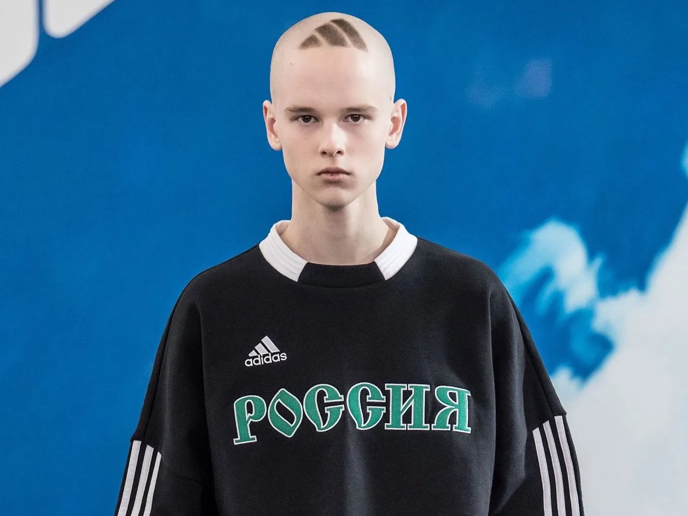 Portrait of a model facing forward against a bright blue backdrop, wearing an oversized black adidas sweatshirt with white stripe detailing on the sleeves and bold green “РОССИЯ” lettering across the chest. The model has a shaved head with geometric hair patterns etched on top, maintaining a neutral expression that complements the stark, graphic styling of the look