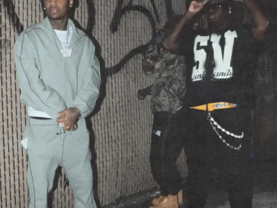 21 lil Harold left side and babydrill right side in a narrow, graffiti-marked alley under harsh flash lighting. On the left, one wears a muted green tracksuit with a matching cap, layered diamond chains, and white sneakers, standing with hands clasped and looking off-frame. On the right, another artist in a black graphic T-shirt, dark jeans, and sunglasses adjusts his cap, accessorized with a bold belt and hanging chain details. A third figure in camouflage and tan boots stands partially turned away in the background. The image carries a gritty, street-style aesthetic, with a “Parental Advisory: Explicit Content” label centered at the bottom