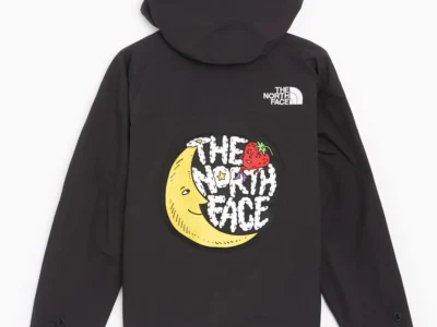 A rear view of a black hooded rain jacket from The North Face, maintaining the same relaxed, utility-driven silhouette seen from the front. The hood sits full and structured at the top, with a clean back panel that emphasizes the garment’s technical construction and minimal seam interruptions. Positioned high on the right shoulder blade is the classic white The North Face, subtly reinforcing brand identity without overpowering the design. The focal point, however, sits at the center of the back: a large, circular graphic patch that introduces a playful, collaborative tone. The graphic features a stylized crescent moon with a face, rendered in a soft yellow tone, paired with a red strawberry motif near the top edge. Overlaid text reads “THE NORTH FACE” in a hand-drawn, cloud-like font, giving the piece a whimsical, almost storybook quality. This visual language suggests a crossover into artistic or collaborative territory—likely tied to a project such as Sky High Farm—blending outdoor gear with expressive, community-driven design. Sleeves remain long with adjustable cuff tabs, consistent with weather-ready functionality. The overall composition balances technical performance with a bold, graphic back statement—transforming a standard rain shell into a piece with narrative and visual identity