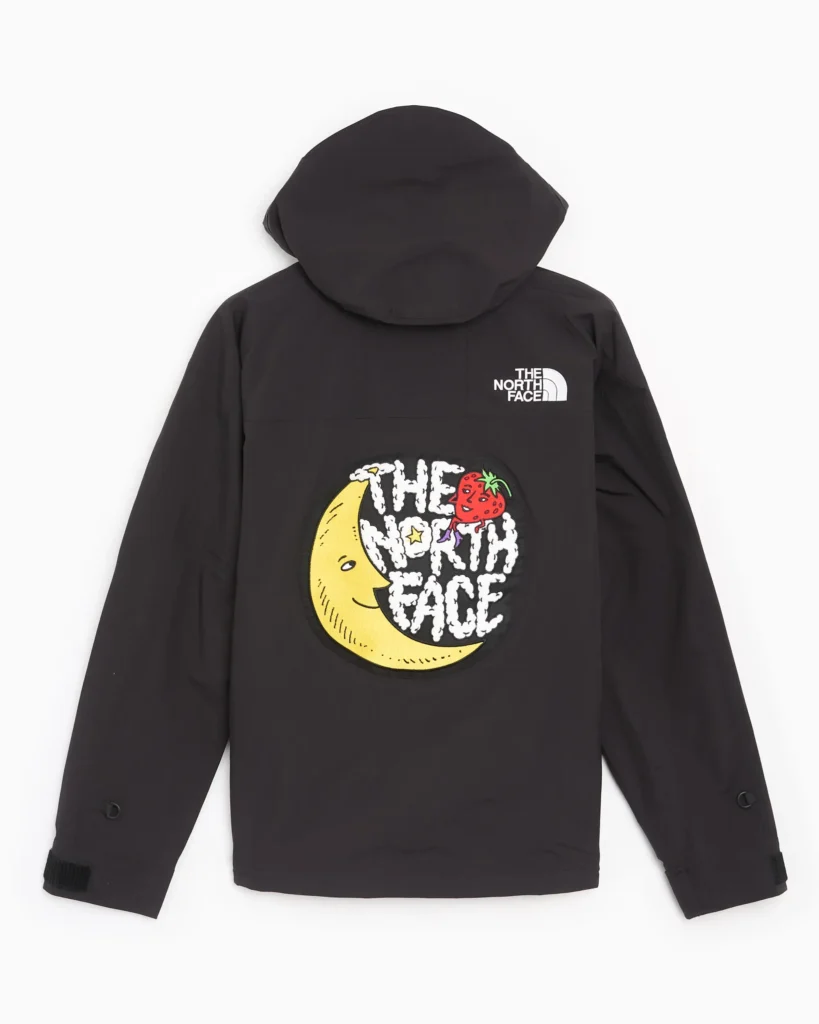 A rear view of a black hooded rain jacket from The North Face, maintaining the same relaxed, utility-driven silhouette seen from the front. The hood sits full and structured at the top, with a clean back panel that emphasizes the garment’s technical construction and minimal seam interruptions. Positioned high on the right shoulder blade is the classic white The North Face, subtly reinforcing brand identity without overpowering the design. The focal point, however, sits at the center of the back: a large, circular graphic patch that introduces a playful, collaborative tone. The graphic features a stylized crescent moon with a face, rendered in a soft yellow tone, paired with a red strawberry motif near the top edge. Overlaid text reads “THE NORTH FACE” in a hand-drawn, cloud-like font, giving the piece a whimsical, almost storybook quality. This visual language suggests a crossover into artistic or collaborative territory—likely tied to a project such as Sky High Farm—blending outdoor gear with expressive, community-driven design. Sleeves remain long with adjustable cuff tabs, consistent with weather-ready functionality. The overall composition balances technical performance with a bold, graphic back statement—transforming a standard rain shell into a piece with narrative and visual identity