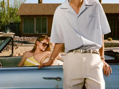 A sun-drenched scene unfolds beside a vintage convertible, where two figures embody a relaxed, mid-century-inflected sense of style and leisure. In the foreground, a young man leans casually against the car door, his posture composed yet effortless. He wears a short-sleeve, open-collar shirt in a pale, airy tone—lightly textured and fluid—tucked into tailored cream trousers secured with a patterned belt. The silhouette is clean and elongated, balancing softness with precision. Dark sunglasses and a fine chain necklace complete the look, adding a subtle, modern sharpness. Behind him, a woman reclines in the passenger seat, partially shaded, her pose languid and unguarded. She wears a yellow striped top and sunglasses, her hand resting near her hair as if caught mid-gesture. Her presence introduces a quiet narrative contrast—less posed, more instinctive—softening the composition. The setting is distinctly desert-modern: a low, wood-toned house sits in the background, framed by palm trees, stone landscaping, and clear blue sky. The powder-blue convertible and cream interior echo the palette of the clothing, creating a cohesive visual rhythm of warm neutrals and sun-faded pastels. The image reads as a study in atmosphere as much as style—where tailoring, light, and environment converge. It evokes a kind of cinematic stillness, rooted in ease and continuity, where the moment feels less staged than simply pausd in time.
