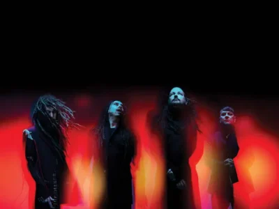 Korn stand in a dark, stylized composition against a blurred red and black backdrop, their figures partially distorted by motion and light. The members wear all-black clothing, their silhouettes elongated and shadowed, evoking tension and unease. The central figure, Jonathan Davis, faces forward with a solemn, almost confrontational presence, while the others angle outward, creating a fragmented, atmospheric formation that reflects the band’s raw and immersive aesthetic