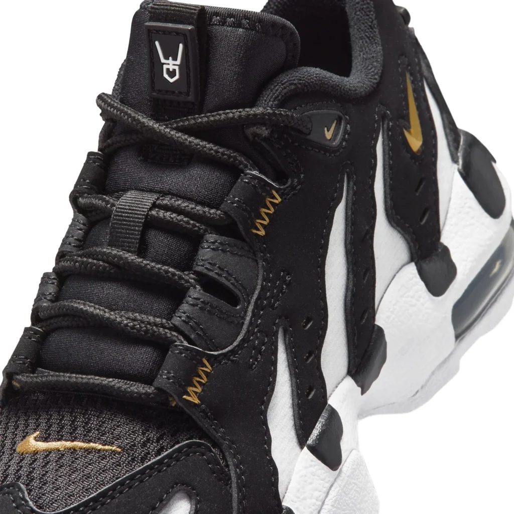 Close-up view of a Nike shoe upper, highlighting black mesh and synthetic overlays with sculpted, wave-like panels over a white base. Black rope laces weave through reinforced eyelets, accented by small gold stitch details. A metallic gold Swoosh appears near the toe and side panel, while the padded tongue features a rubberized logo patch at the top. The textured white midsole and visible Air unit edge are partially visible along the side