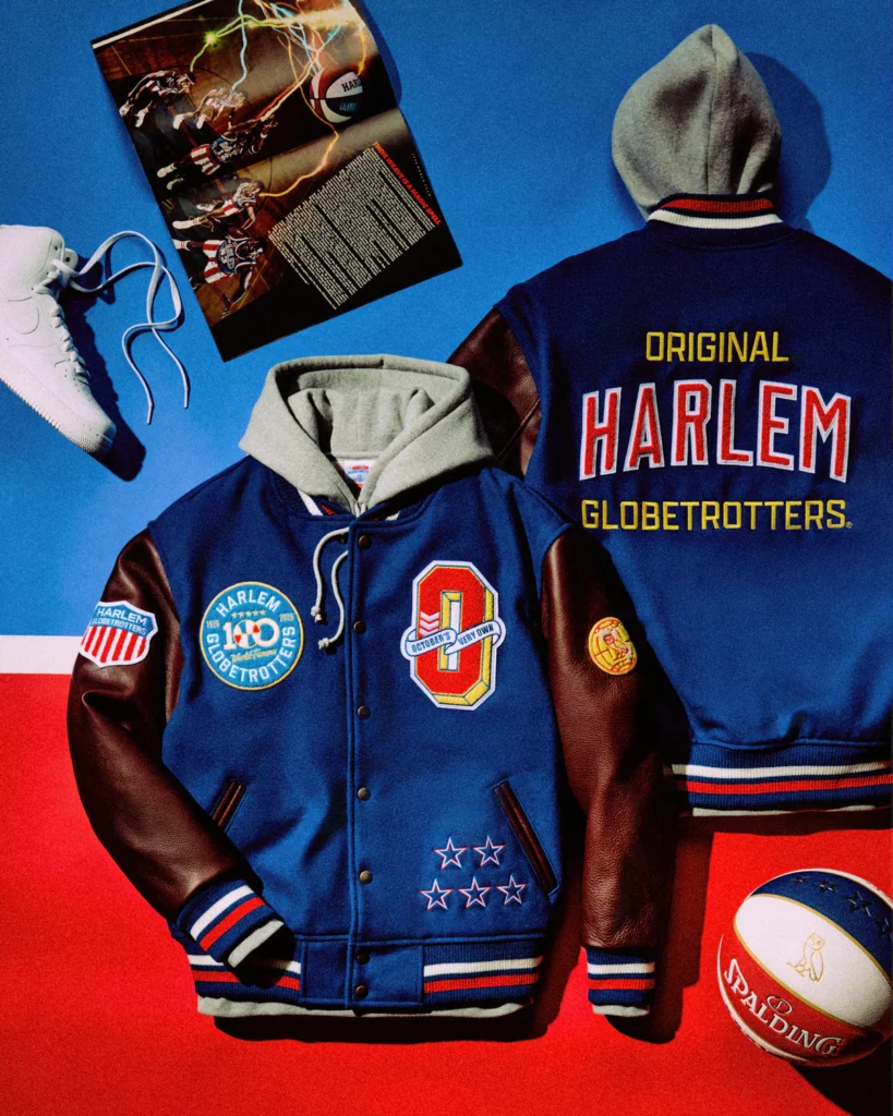OVO x Harlem Globetrotters varsity jacket in blue with leather sleeves and gray hood, featuring embroidered centennial patches and “Original Harlem Globetrotters” text on the back, styled with a Spalding basketball, white sneakers, and vintage basketball magazine imagery