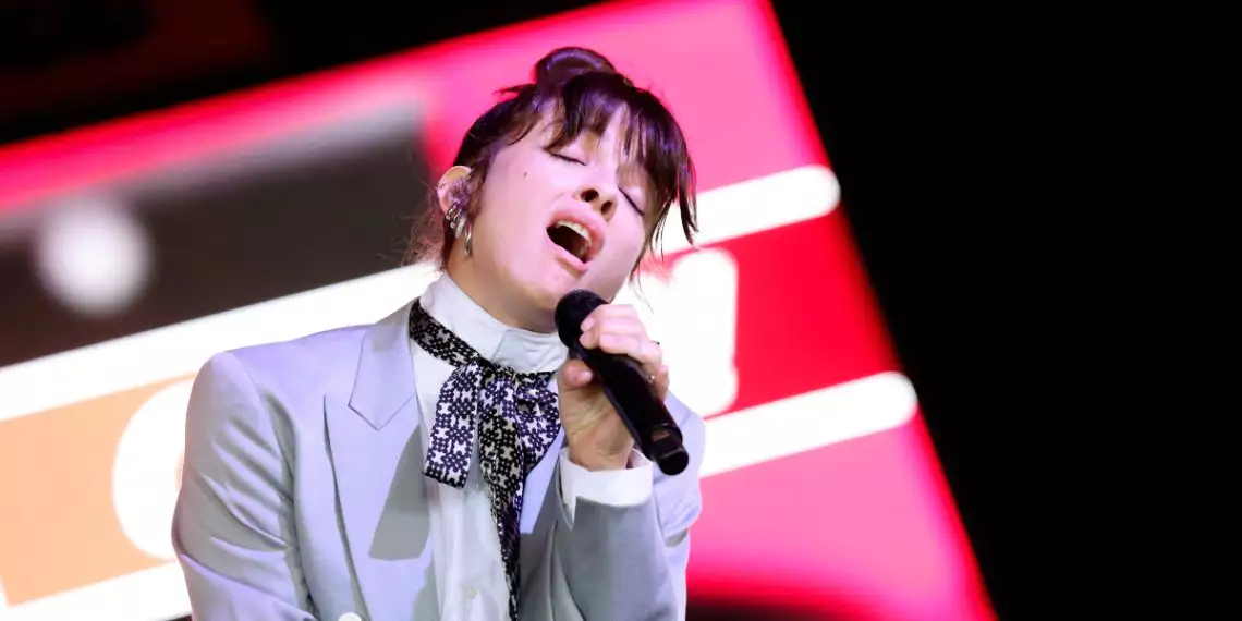 Noga Erez performing on stage with eyes closed mid-vocal, holding a microphone while dressed in a tailored light suit with a patterned scarf, set against bold red and white stage view