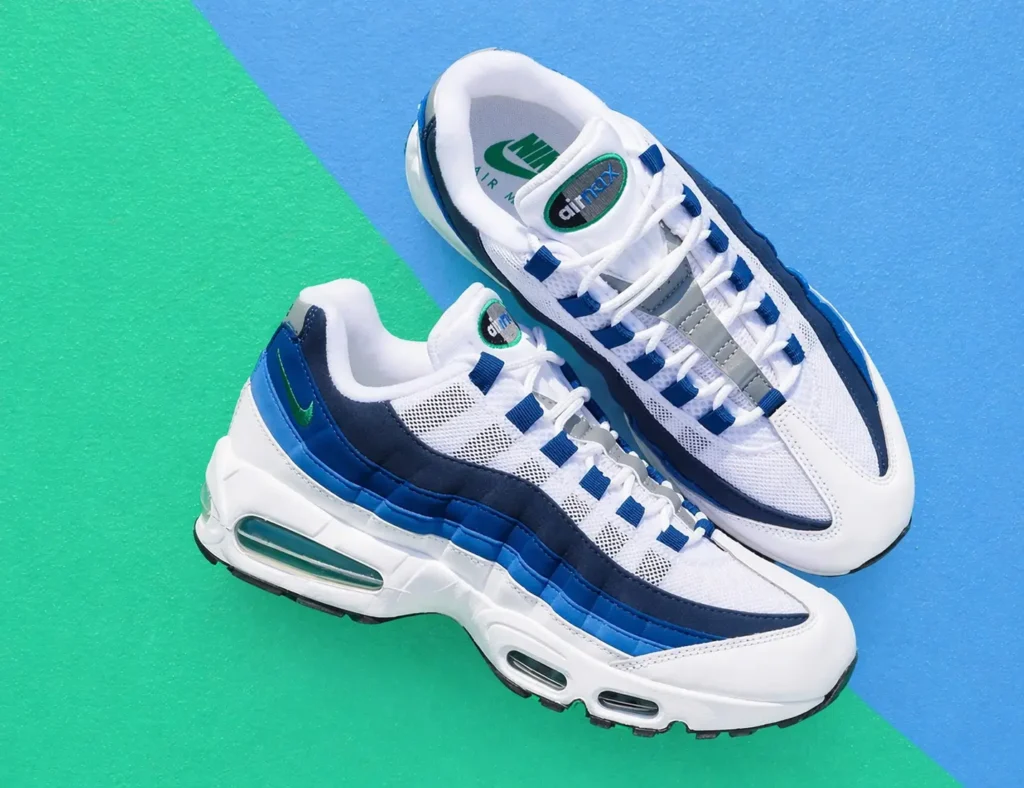A pair of Nike Air Max 95 shoe in white with navy and blue gradient overlays, arranged diagonally on a split green and blue textured surface, featuring visible Air units and mesh paneling in bright