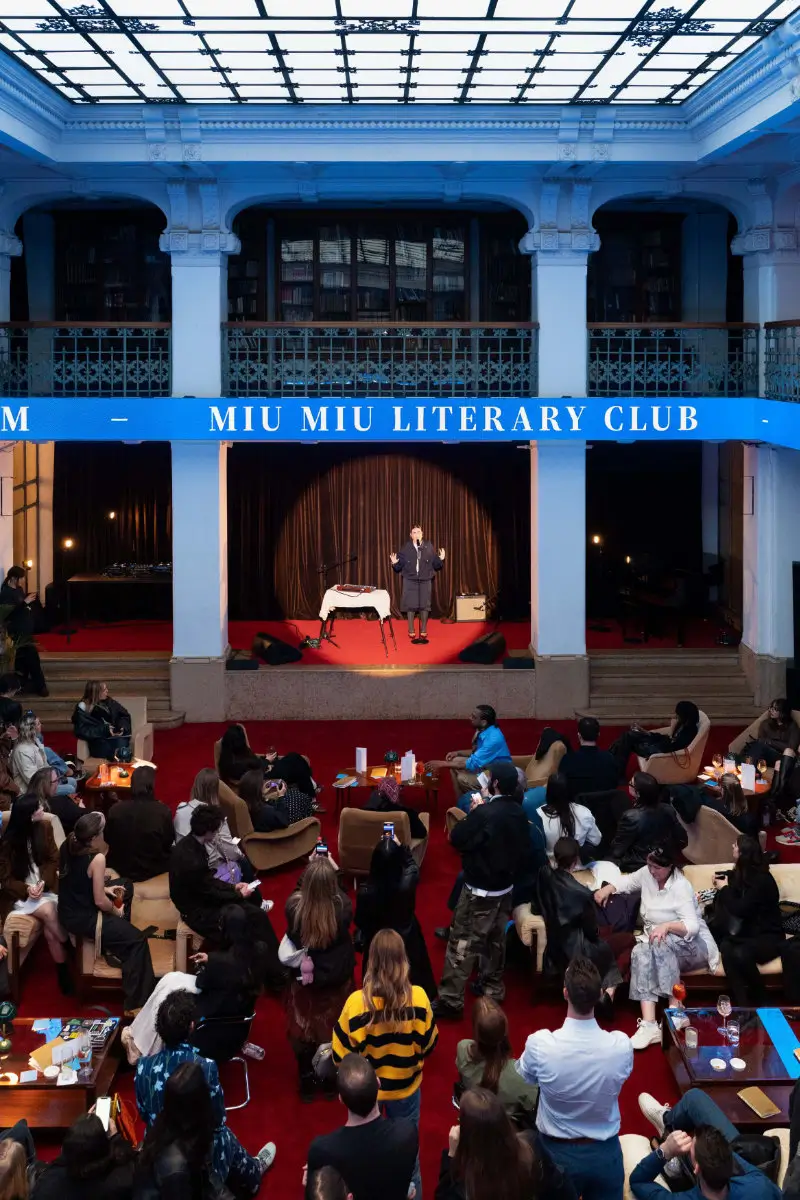 Miu Miu Literary Club live event at Circolo Filologico Milanese featuring a speaker on a small stage with red carpeting beneath a blue “Miu Miu Literary Club” banner, surrounded by a seated audience in an intimate, salon-style setting, blending literature, performance, and fashion within a historic architectural interior