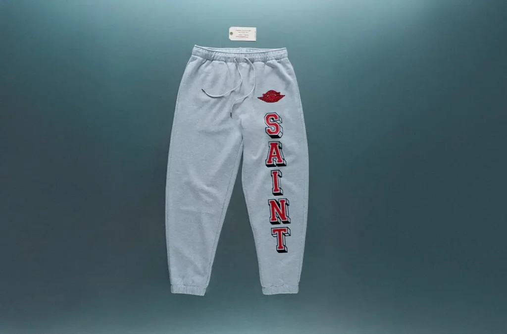 Light grey SAINT Mxxxxxx x Jordan Brand sweatpants with vertical “SAINT” lettering in red and black down the leg, featuring an embroidered red Jumpman-style wing logo on the upper thigh and elastic waistband with drawstring, displayed against a muted blue wall backdrop
