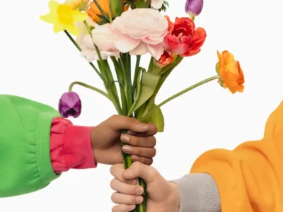 Close-up of two hands exchanging a colorful bouquet of mixed flowers against a plain white background. One hand, wearing a bright green sleeve with a pink cuff, passes the bouquet to another hand in an orange sleeve. The arrangement includes tulips, daffodils, and other spring blooms in vivid shades of yellow, purple, red, and soft pink. Subtle text at the bottom reads “ГОША РУБЧИНСКИЙ” and “gosharubchinskiy.com.”