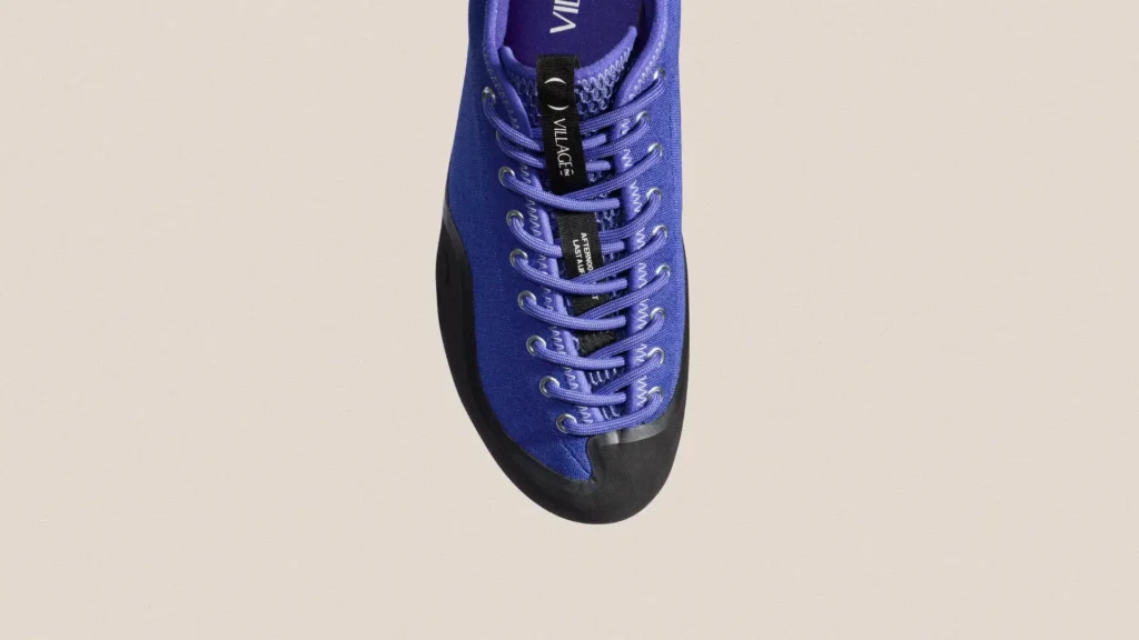 Top-down view of VILLAGE PM 1PM sneaker in natural purple canvas with black rubber toe cap, tonal purple laces threaded through metal eyelets, and central black pull tab with VILLAGE branding against a neutral background