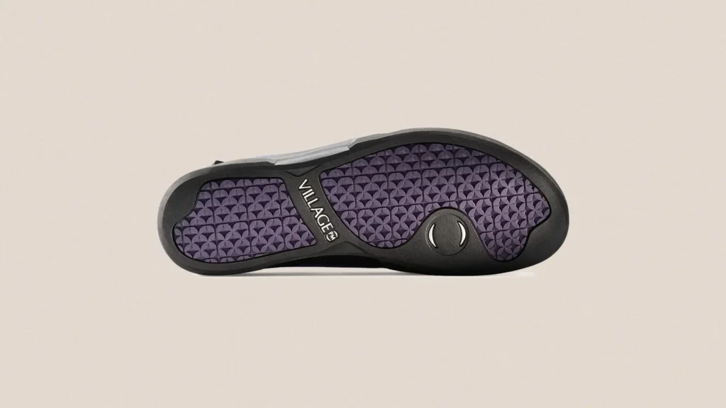 Outsole view of VILLAGE PM 1PM sneaker featuring a black rubber frame with textured purple traction pattern, central VILLAGE branding, and circular grip detail, displayed against a neutral background