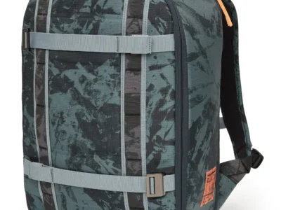 A three-quarter front view of a structured teal-green backpack with a tonal camo-style pattern, emphasizing its rounded, semi-rigid silhouette. The front panel is organized with vertical webbing columns intersected by two horizontal compression straps, each secured with metal adjusters, reinforcing its modular, utility-driven design. Subtle orange accents appear along the top zipper and pull tabs, adding contrast to the otherwise muted palette. The side profile reveals a clean, slightly tapered structure with a secondary zip compartment and a small leather patch detail near the base, introducing a refined branding element. The materials appear durable and lightly textured, suggesting water resistance and everyday resilience. Overall, the design balances technical function with a contemporary urban aesthetic, positioned between outdoor gear and city-ready carry